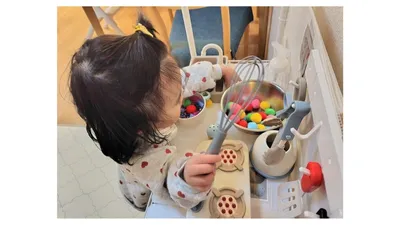 おもちゃのキッチンで、泡立て器を持って混ぜるまねをする女の子